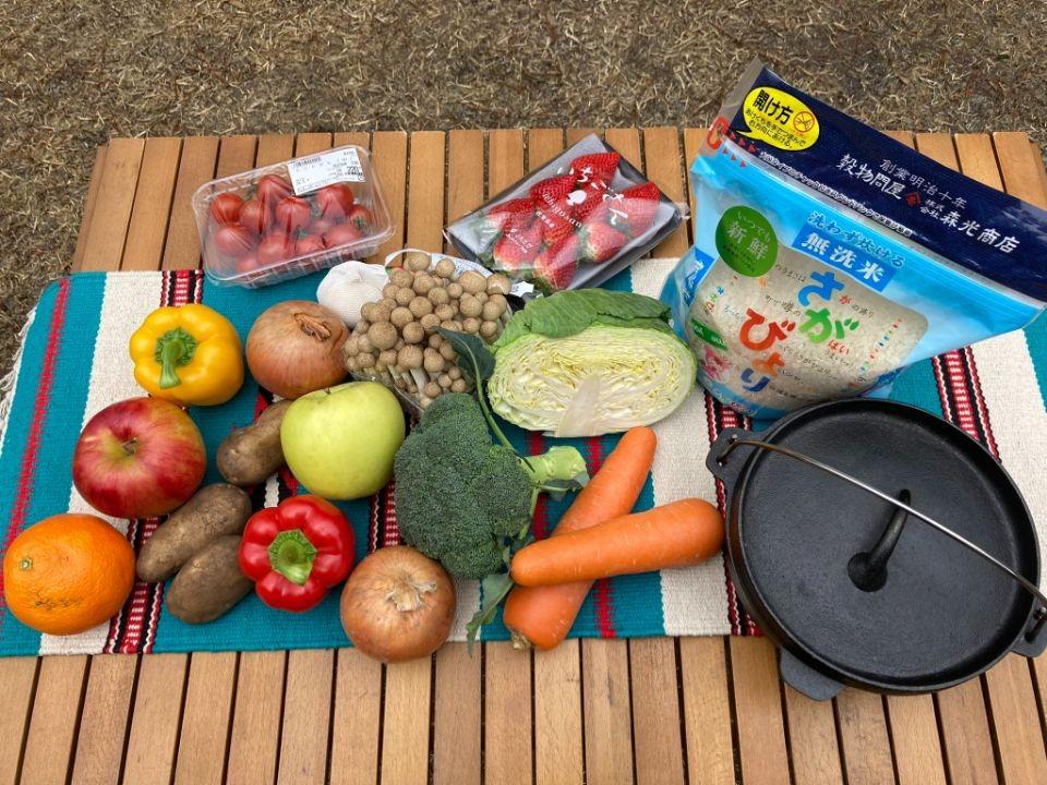 写真：折り畳みテーブルの上に食材が並べられた様子。右端に鉄鍋と米2キログラムの袋、中央に大量の野菜、奥にいちご、左端にりんごがある