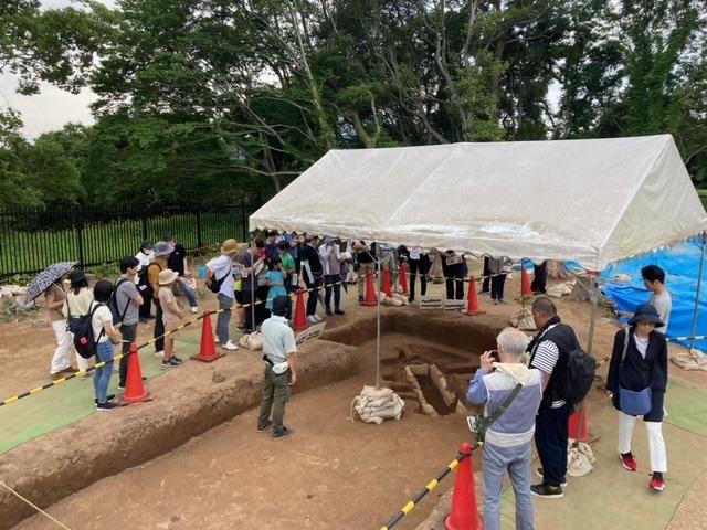 写真：発掘された石棺墓の上にテントが張られ、20～30人の見学者が石棺墓を取り囲んでいる