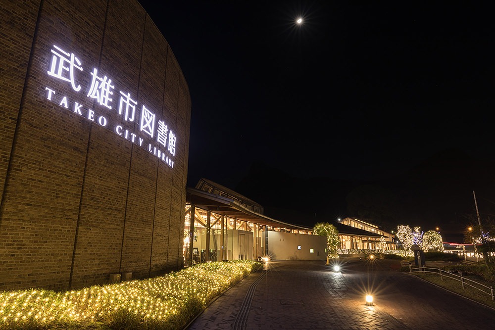 写真：武雄市図書館の外観。「武雄市図書館」の電飾看板が点灯し、建物の周囲の植え込みにイルミネーション装飾が施された夜景