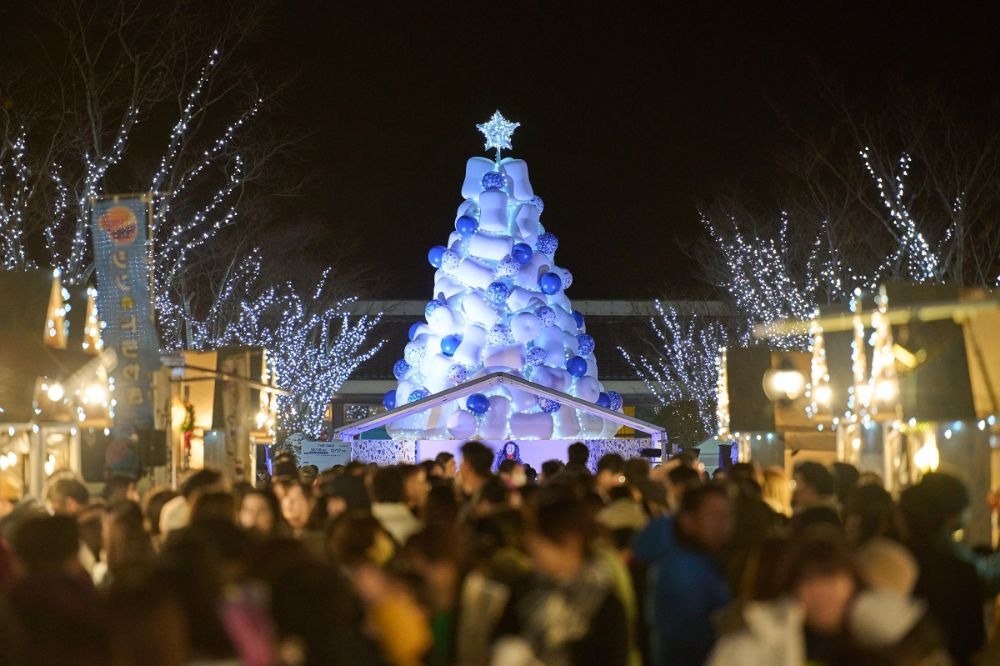 写真：中央に大きなマシュマロを積み重ねたイメージのクリスマスツリーが設置され、多くの来場客でにぎわっている広場の夜景