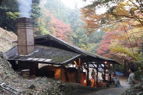 写真：坂の上から登り窯を眺める風景。煙突から煙が上がり、背景の山は紅葉している。登り窯の周りに作業している人が3人ほど見える