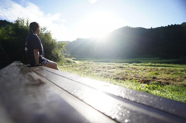 写真：ベンチに腰掛けて風景を眺めている女性。奥の山ぎわから朝日が昇っている