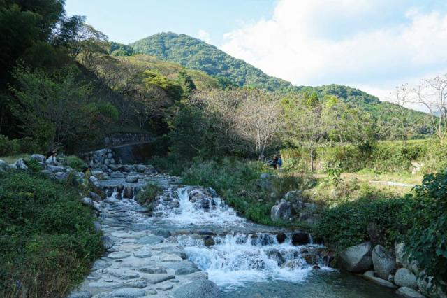 写真：大きな石で護岸された小川。右側の川岸の石に座っている人がいる。左側の川沿い歩道を歩く人が2人いる。背景に山が見える