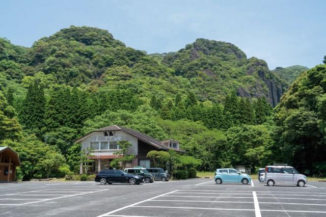 写真：竜門キャンプ場の駐車場。手前に管理棟と駐車車両が7台ほどある。遠景に山が見える