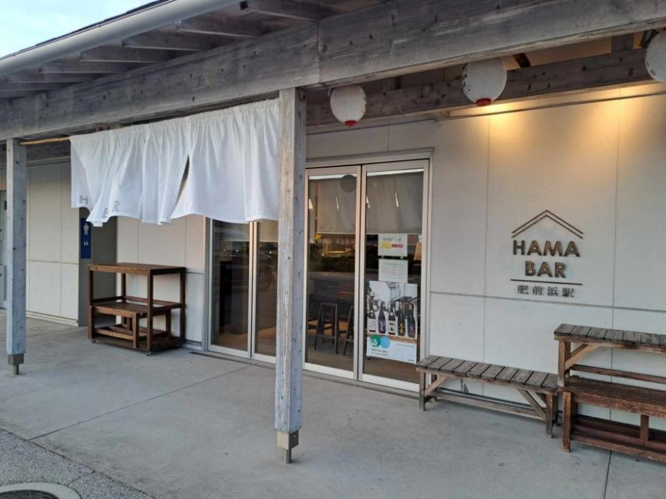 写真：駅舎の外観。「HAMA BAR 肥前浜駅」のロゴサインが壁にかかっている。軒に暖簾、軒下に提灯が3つ並んで下げられている