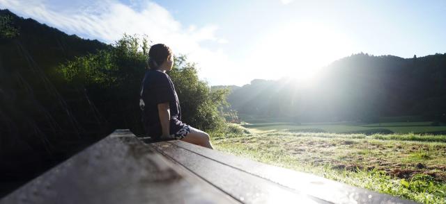 写真：ベンチに腰掛けて風景を眺めている女性。奥の山ぎわから朝日が昇っている