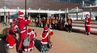 写真：武雄駅前でイベント運営スタッフが挨拶を行っている様子。クリスマスにちなんだ扮装の参加者グループが挨拶している人にカメラを向けている。