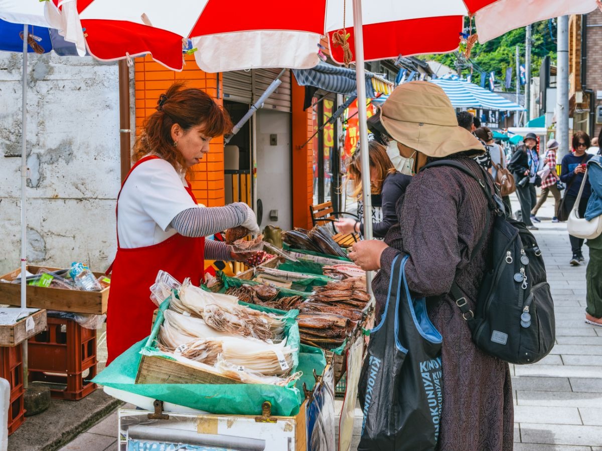 写真：パラソルの下でイカの干物を打っている女性と買い物客2名