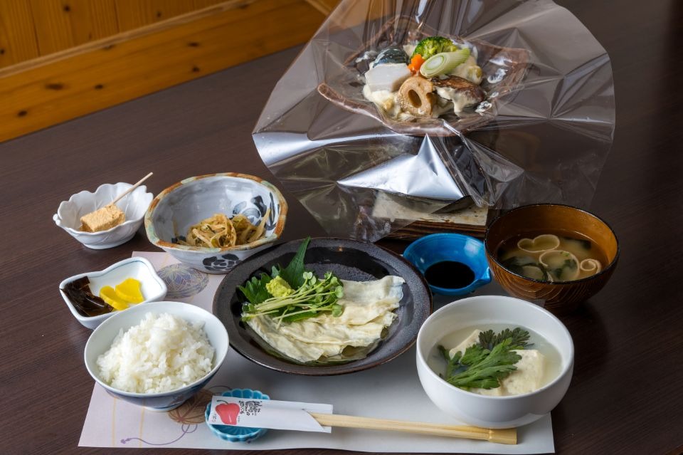 奥に野菜に豆腐ソースをかけた透明フィルムの包み焼と温泉湯豆腐、小鉢、ご飯、味噌汁などが並んだ料理画像